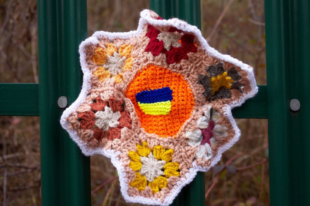 Bright crocheted floral piece with Ukrainian colors on green fence outdoors.