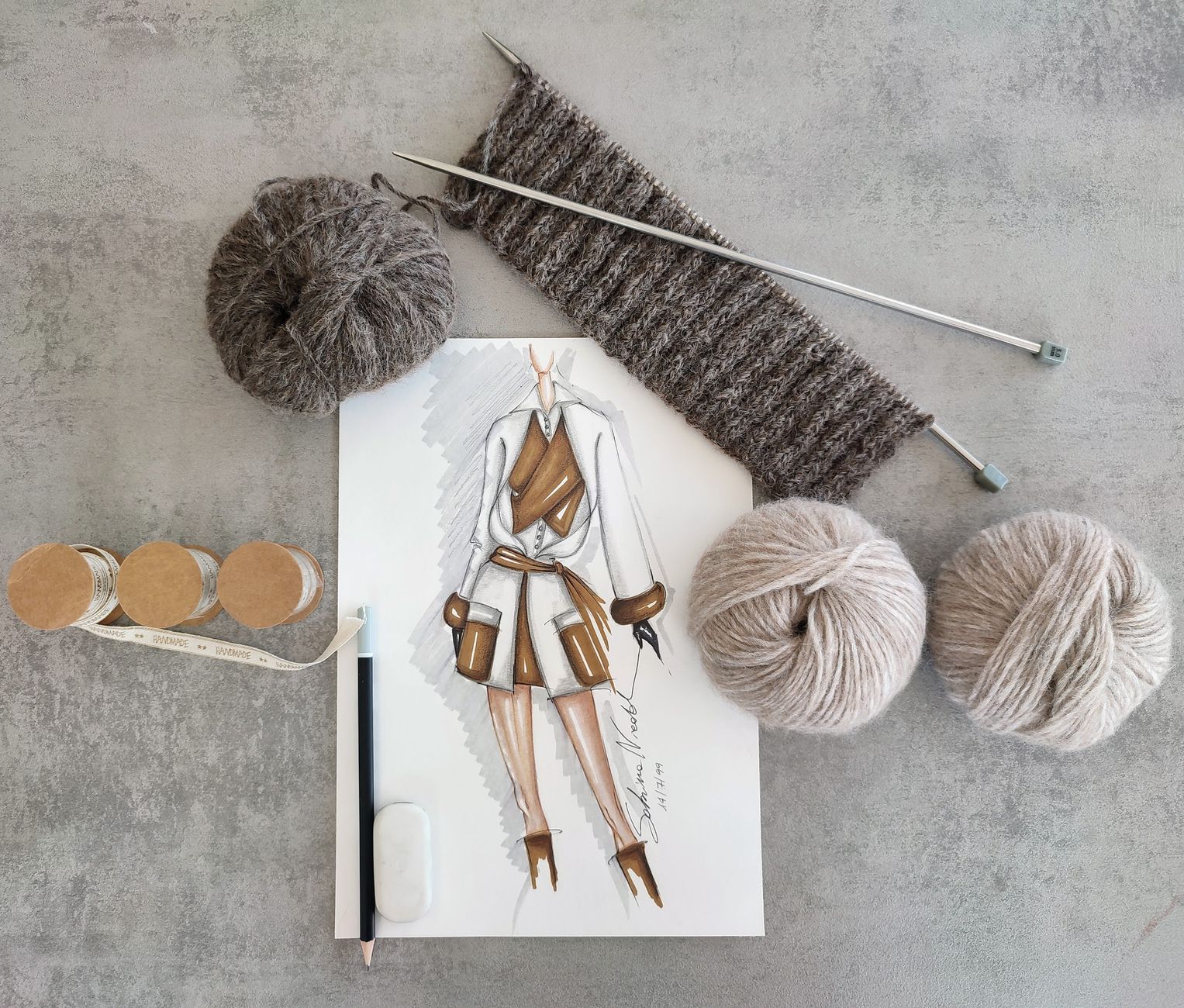 Top view of a fashion sketch surrounded by yarn, needles, and knitting threads on a gray background.