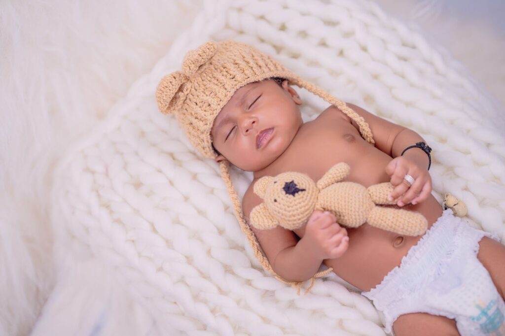 A peaceful baby sleeping on a cozy white blanket, wearing a knitted hat and holding a crocheted bear toy.