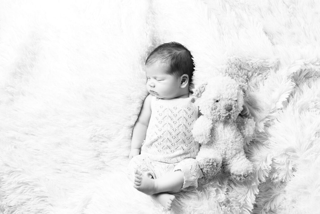 A black and white photo of a sleeping newborn baby cuddling a fluffy teddy bear.