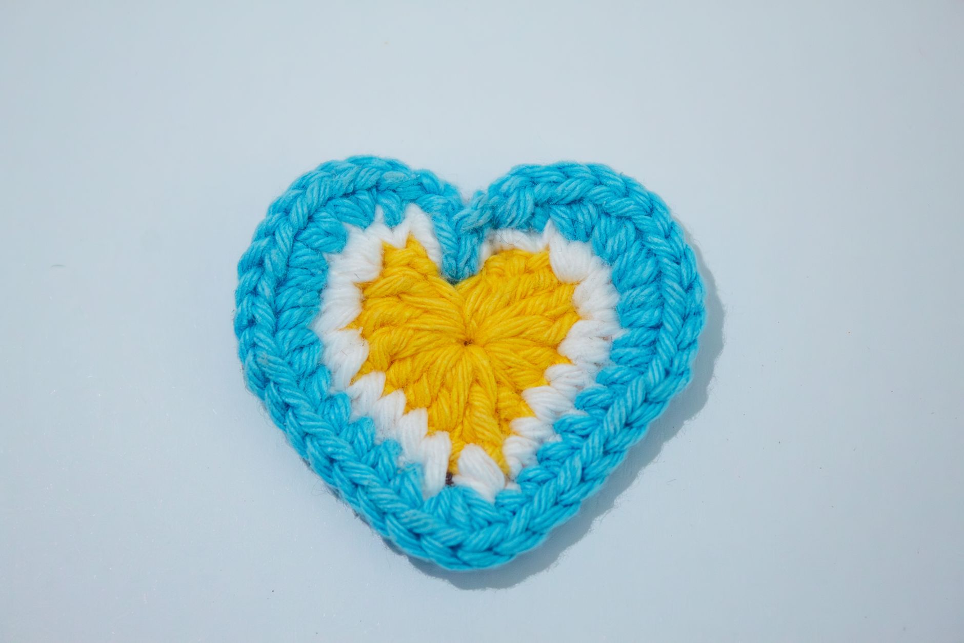 Handmade crocheted heart with blue, white, and yellow yarn, symbolizing love and craft creativity.