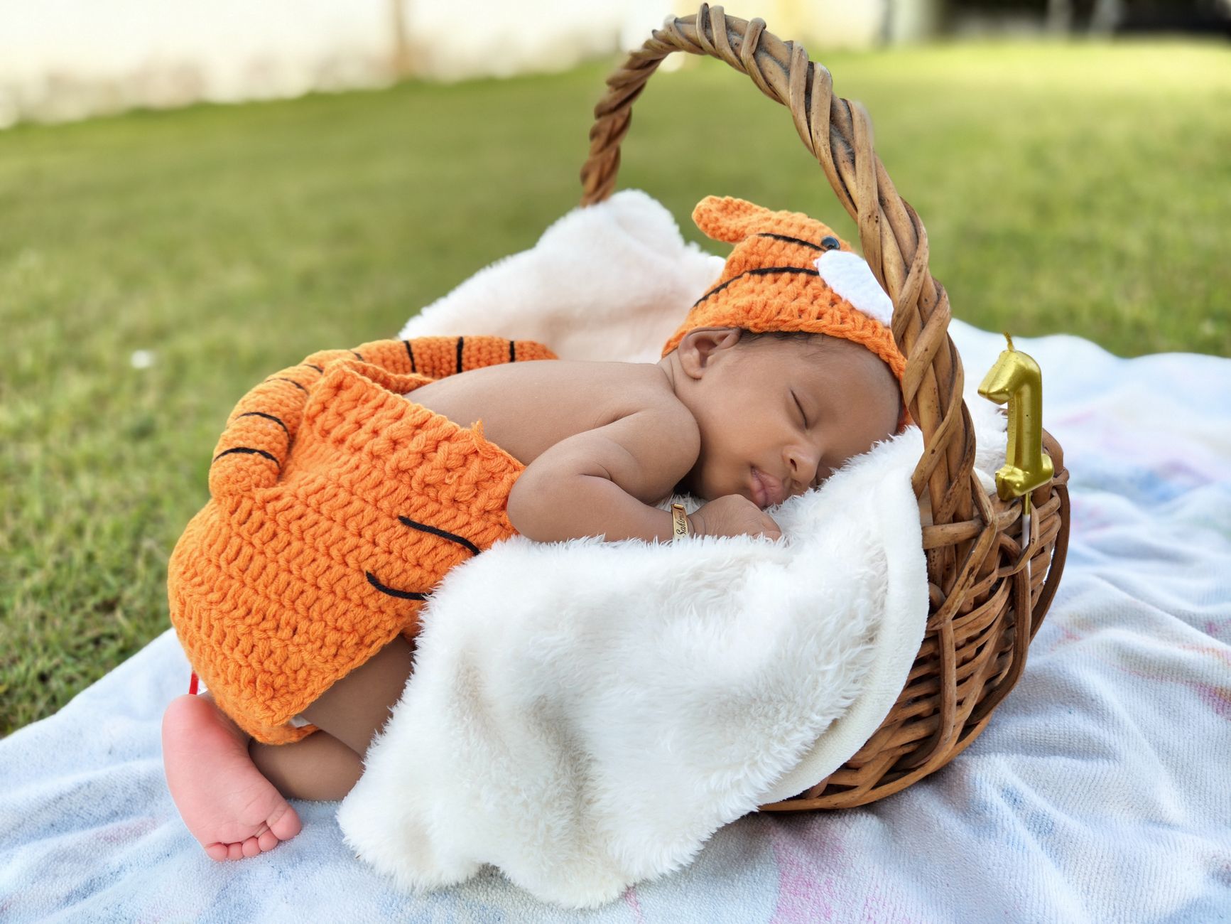 A cute baby in a knitted tiger costume sleeps peacefully in a wicker basket on a sunny day.