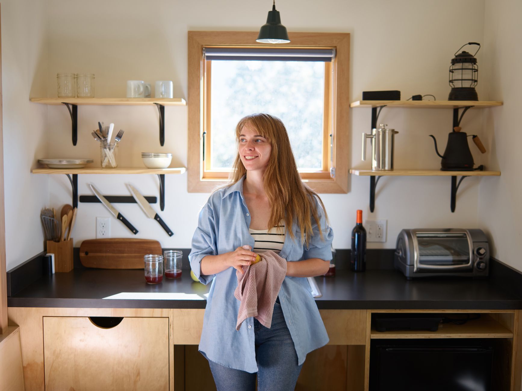 Charming smiling young blonde in casual wear leaning on kitchen counter with dishcloth and apple during sunny morning in contemporary cozy kitchen