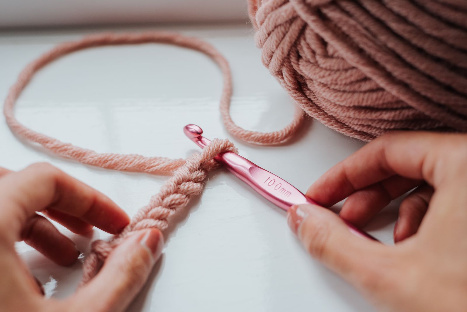 Crop faceless female holding yarn for knitting and pink crochet hook while working on windowsill at home