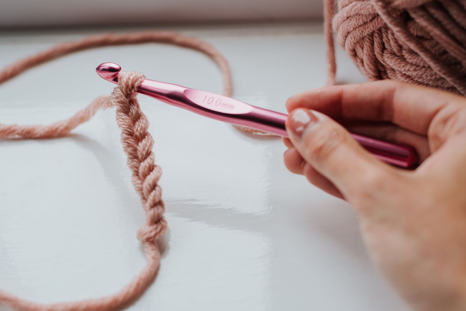 Unrecognizable person using beige threads and pink glossy crochet needle while knitting row of loops on windowsill in daylight