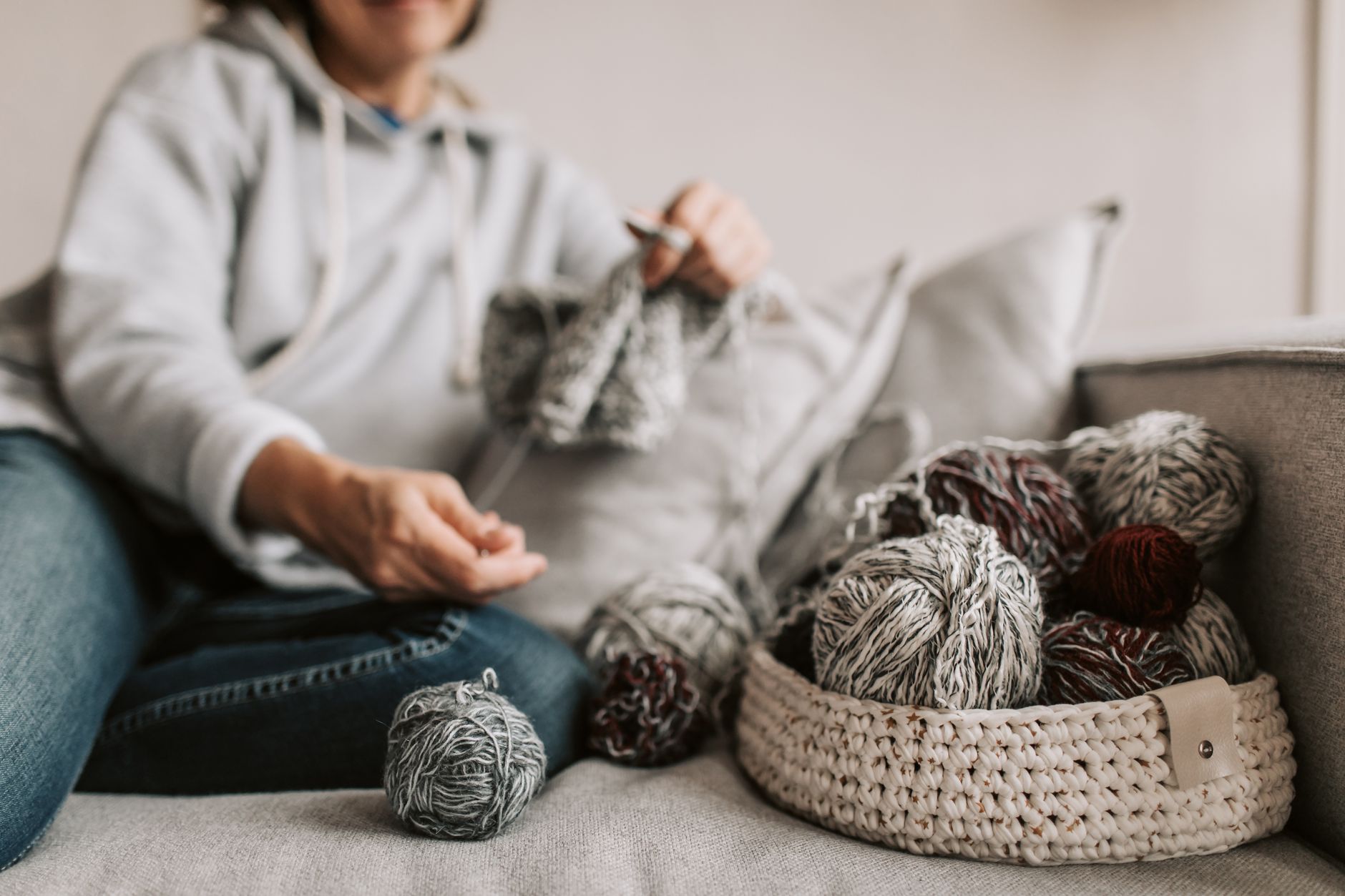 A woman knitting on a couch surrounded by yarn balls in a woven basket.
