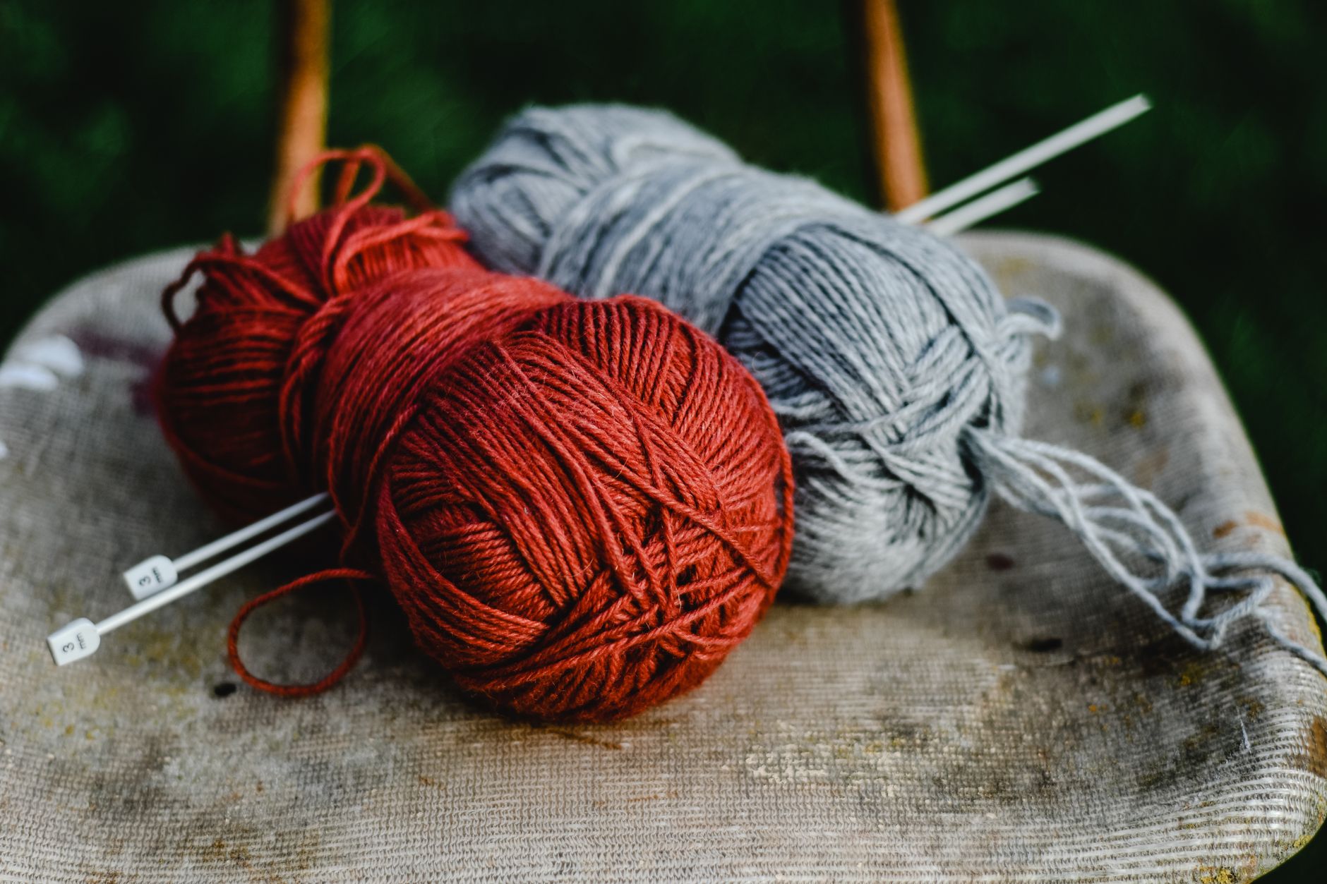 Aesthetic close-up of red and gray yarn balls with knitting needles on a rustic chair.