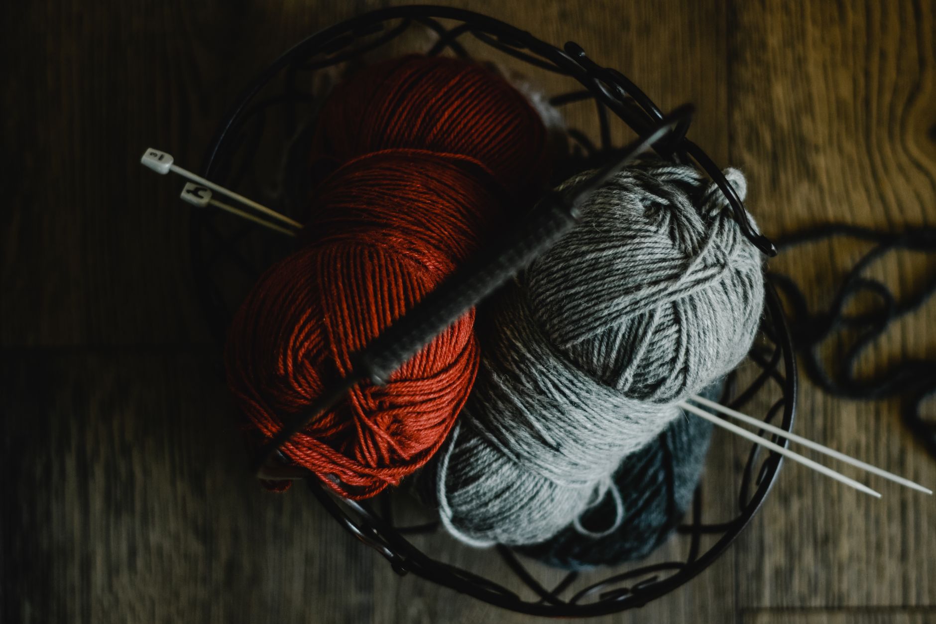 Basket filled with red and gray yarn balls with knitting needles on wooden floor.