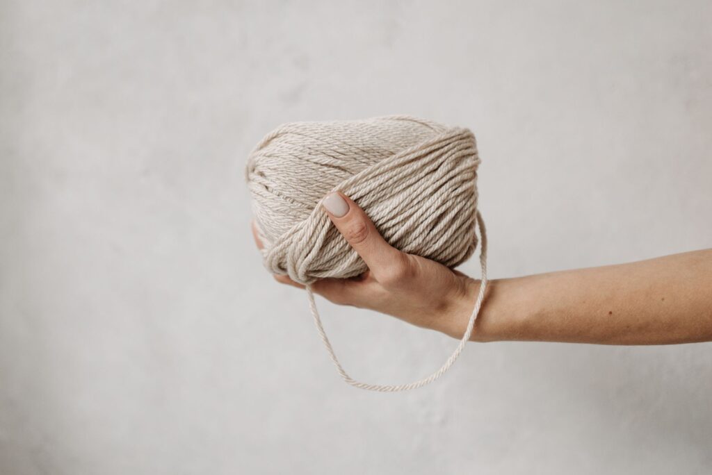 A close-up of a hand gracefully holding a soft beige yarn ball against a neutral background.