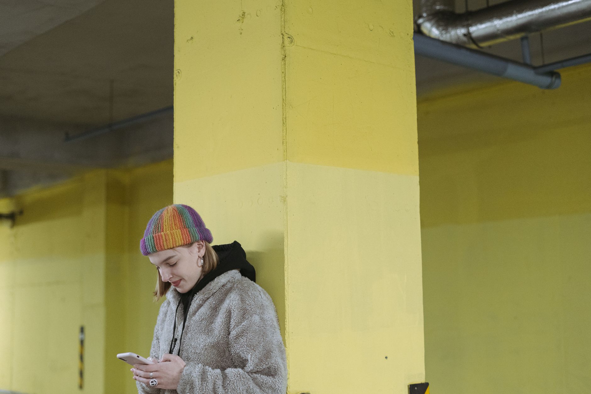 A woman leans against a yellow column while using her smartphone in an urban environment.