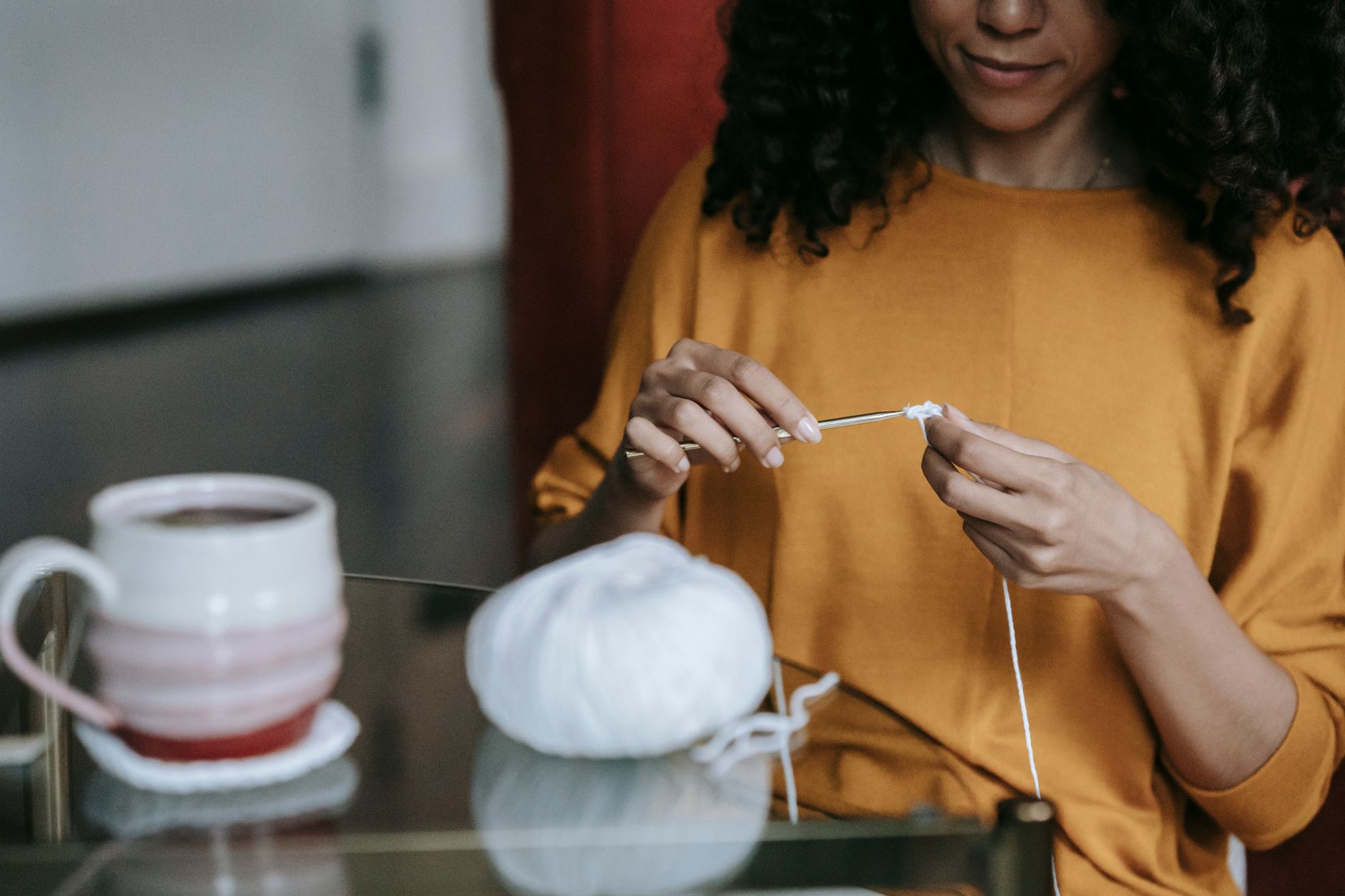 A woman focuses on her crochet work with a coffee mug beside her. Indoor setting, warm ambiance.
