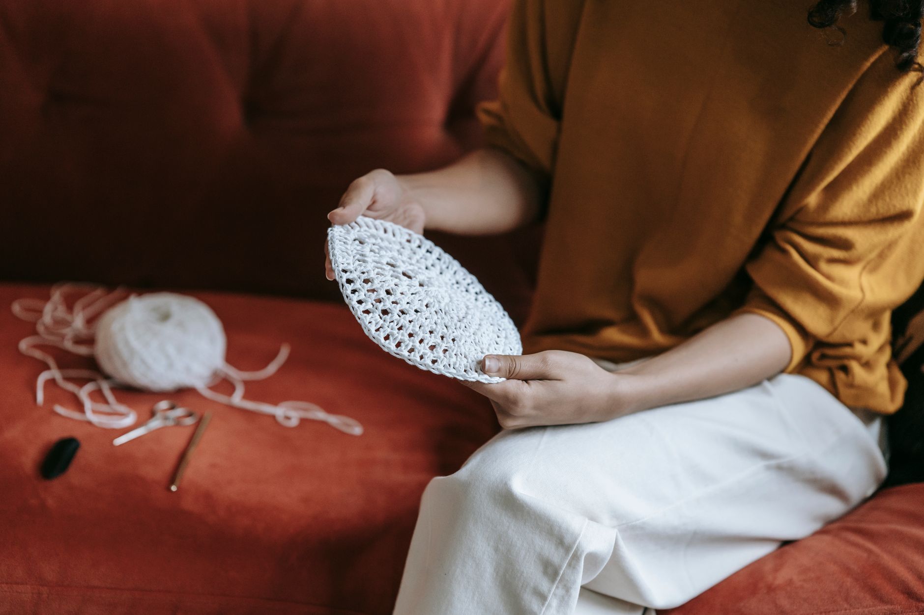 A woman sits on a red sofa crochet stitching a white pattern, evoking warmth and creativity.