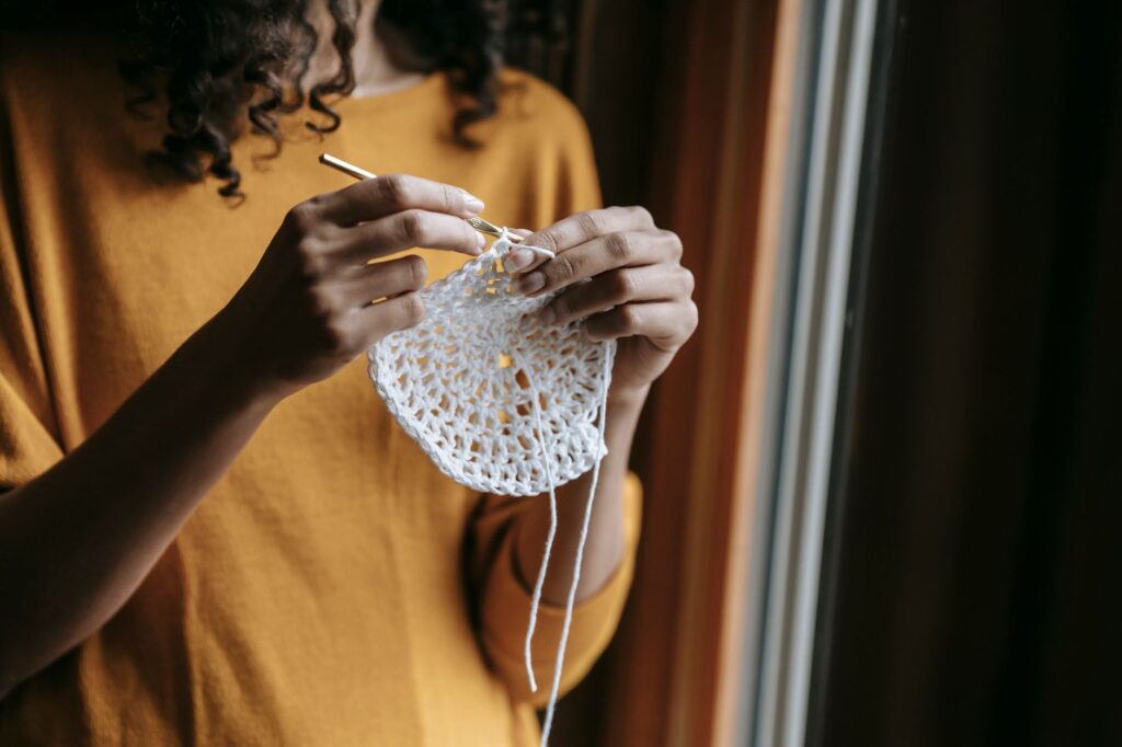 Crop anonymous female artisan with yarn and hook crocheting while having spare time in house